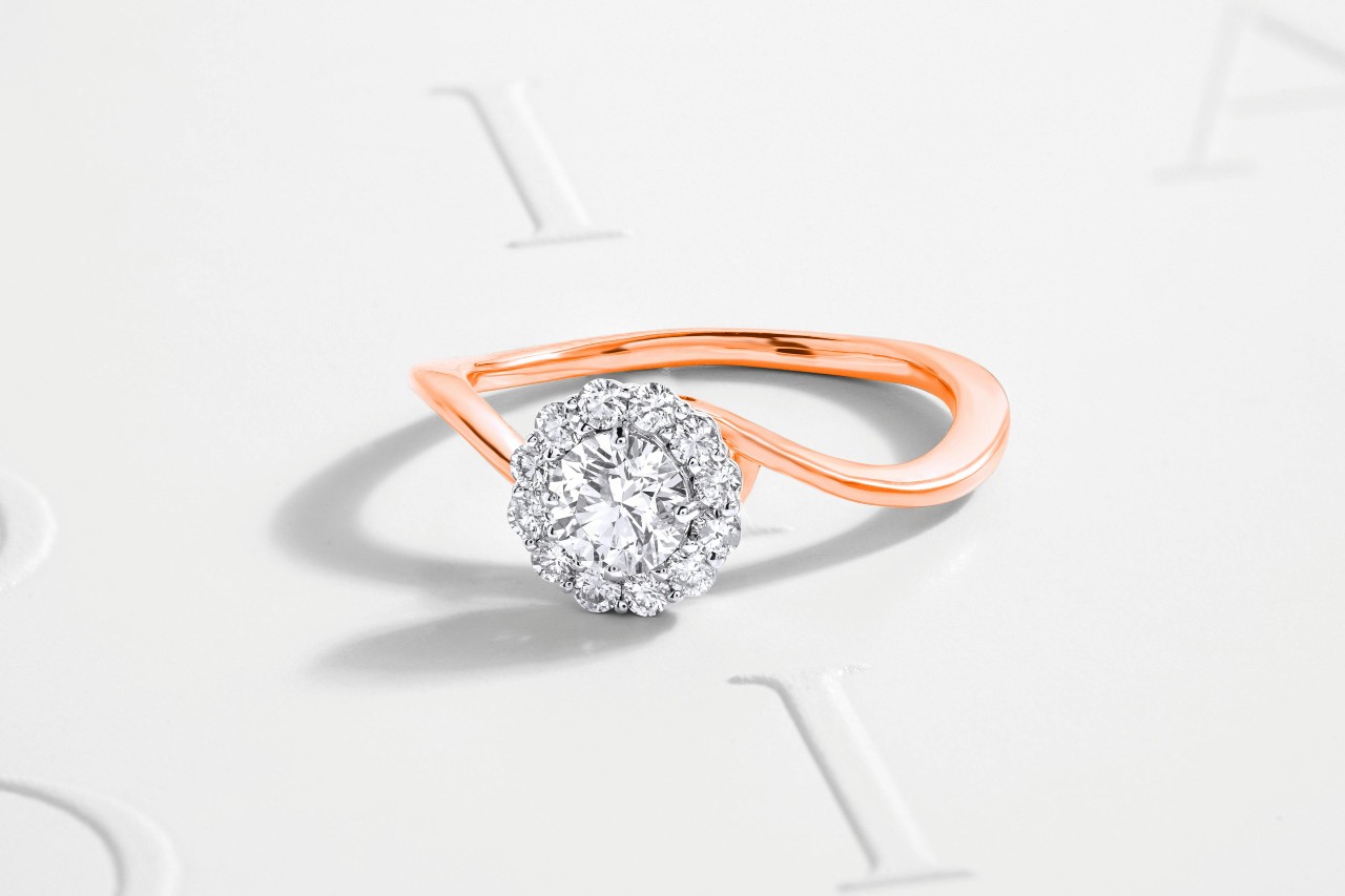 A rose gold engagement ring sits on a paper with embossed letters.