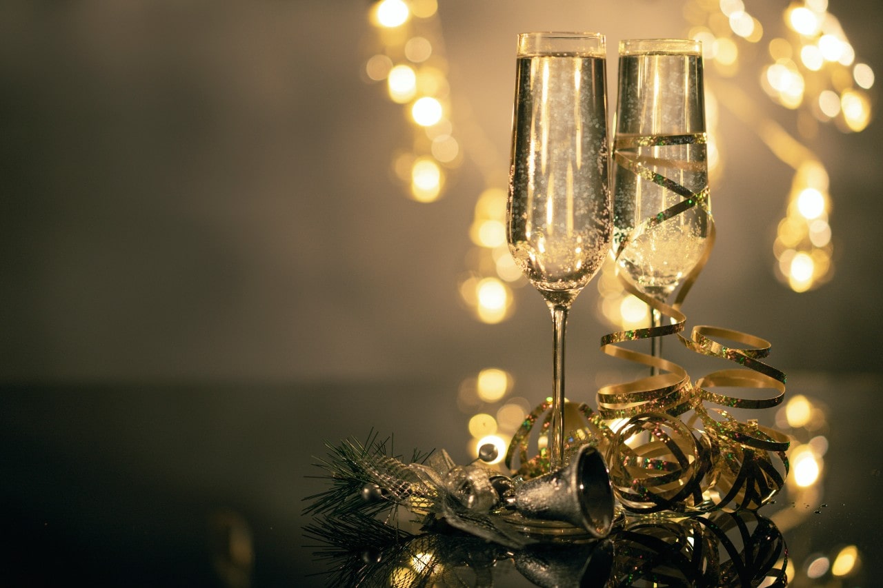 Two champagne flutes sit on a reflective surface with gold ribbon and decorations.