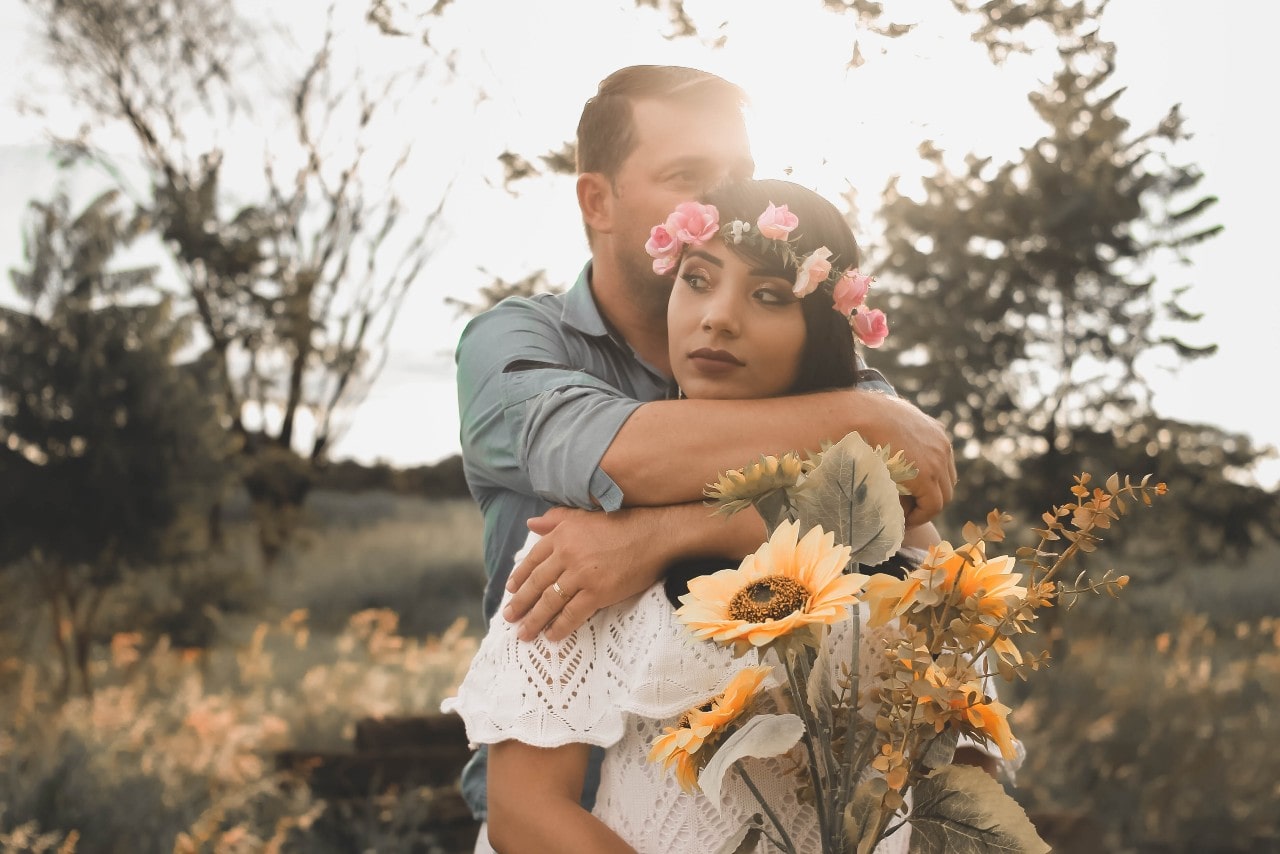 A groom embracing his bride, holding a bouquet of sunflowers and eucalyptus, from behind.