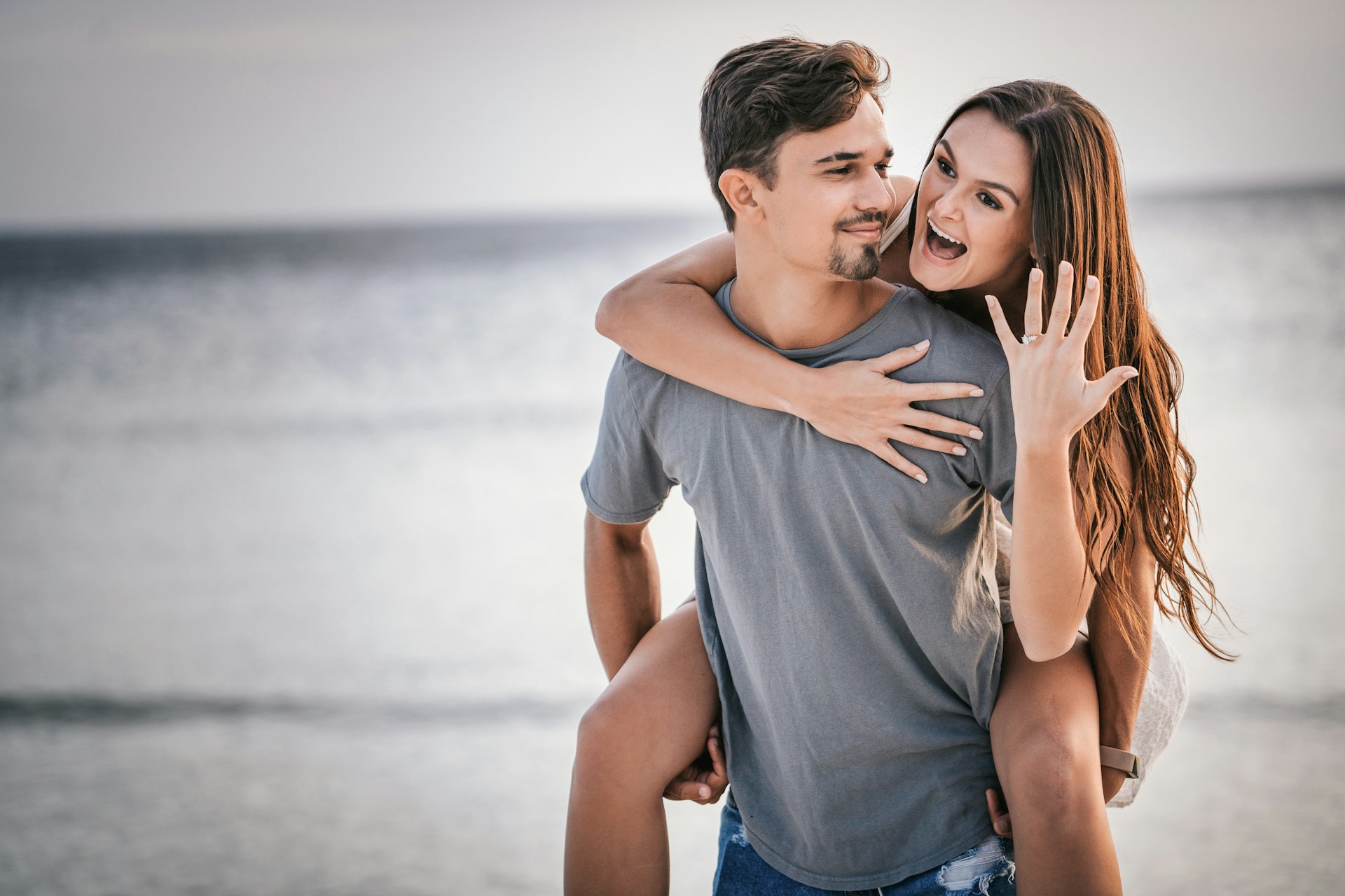 lady riding on a man’s back on the beach smiling with an engagement ring