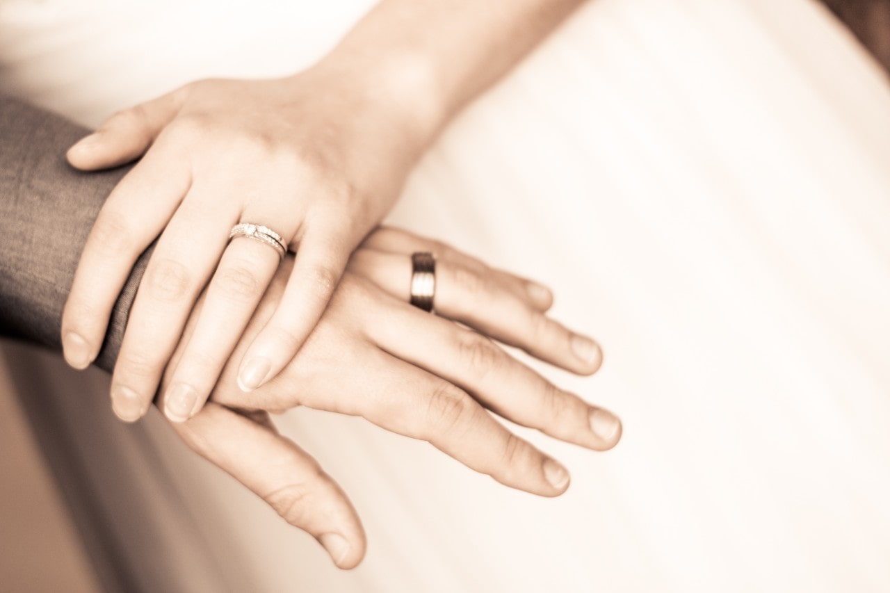 A woman’s hand rests on a man’s, both wearing wedding bands.